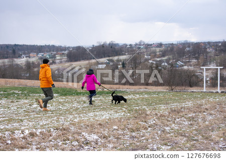 Couple strolls through a snowy field with their dog on a chilly winter day while enjoying the scenic views of distant houses and nature 127676698