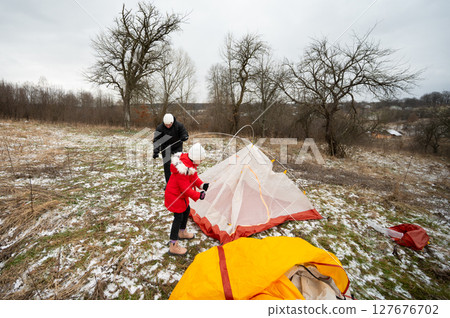 Setting up a colorful tent on a chilly winter day while enjoying time outdoors with a child in a serene countryside landscape Setting up a colorful tent on a chilly winter day while enjoying time outdoors with a child in a serene countryside landscape 127676702