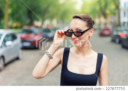 Confident woman in black dress lowers sunglasses with playful expression while standing on city street. Elegant and stylish look. Female person walking city street Confident woman in black dress lowers sunglasses with playful expression while standing on city street. Elegant and stylish look. Female person walking city street 127677753