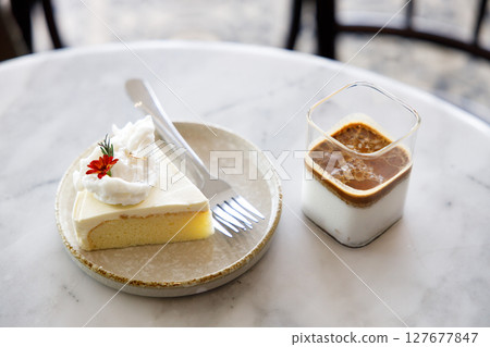 Cup of milk dirty coffee with a piece of coconut cake on white marble table by the window. Cup of milk dirty coffee with a piece of coconut cake on white marble table by the window. 127677847