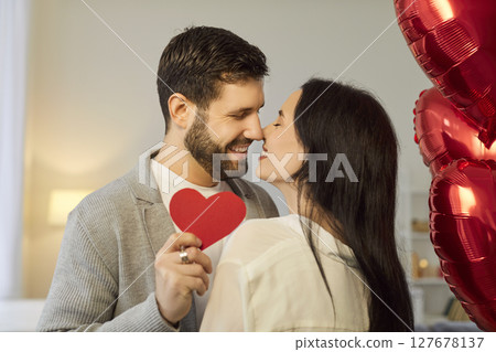 Happy young couple hugging, kissing, holding red paper heart during romantic Valentine Day at home. Happy young couple hugging, kissing, holding red paper heart during romantic Valentine Day at home. 127678137