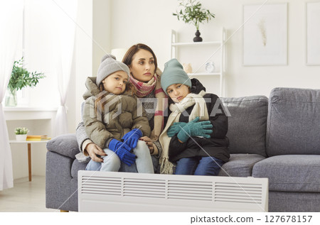 Mother with kids sitting on sofa in winter outwear at home and trying to warm with electric heater. Mother with kids sitting on sofa in winter outwear at home and trying to warm with electric heater. 127678157