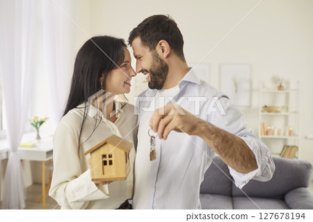 Happy young couple holding keys and toy house, smiling joyfully, celebrating buying their new home. 127678194