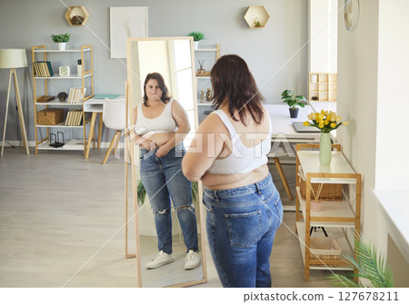 Fat overweight woman looking in the mirror standing in living room at home. 127678211