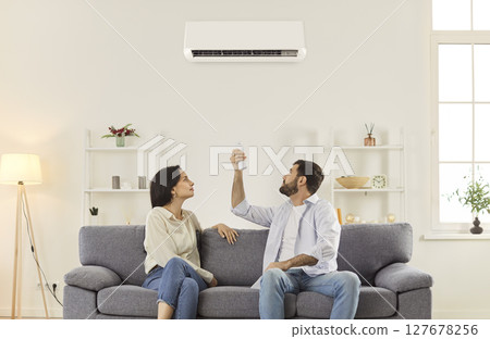 Young couple sitting on sofa in living room, turning on air conditioner with remote controller. 127678256