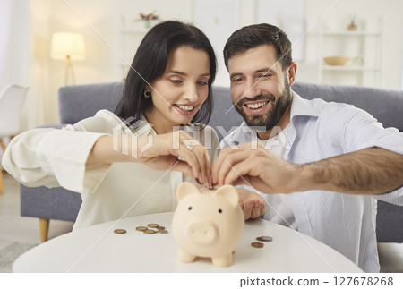 Happy young couple sitting by desk, putting coins into piggy bank, planning finances and future. Happy young couple sitting by desk, putting coins into piggy bank, planning finances and future. 127678268