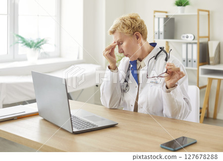 Tired female doctor resting at desk, putting off glasses and rubbing nose after working on laptop. Tired female doctor resting at desk, putting off glasses and rubbing nose after working on laptop. 127678288