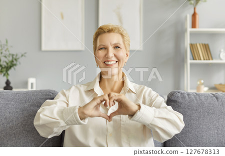 Smiling mature woman with short blonde hair sitting on cozy sofa, forming heart shape with her hands 127678313