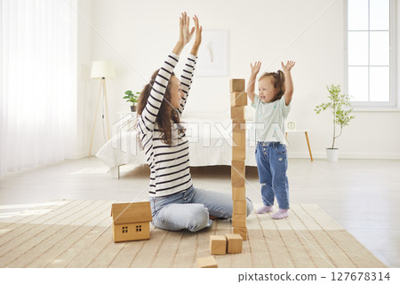 Joyful mother and her young daughter engage in playful activity, building tower with wooden blocks. 127678314