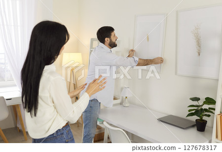 Joyful young couple decorating their new home together, hanging picture on wall, working as team. 127678317