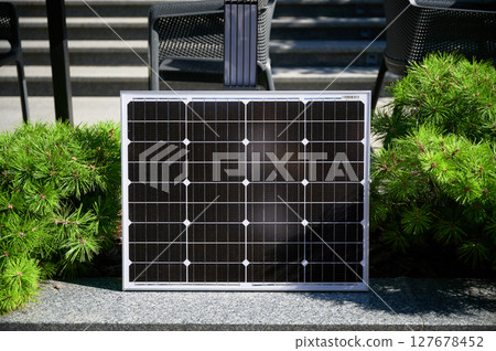 Photovoltaic solar panel against backdrop of vibrant green shrubs on stone ledge, soaking up sunlight in modern, urban landscape. Concept of integration of renewable energy within city environments. 127678452
