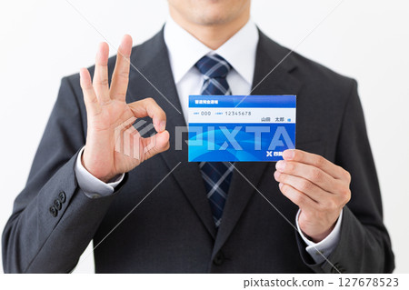 A middle-aged male businessman wearing a suit holding a bank deposit book and making a circle sign 127678523