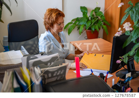 Woman working with folders and documents at her desk in an office setting. Paperwork routine, archive management, and administrative desk tasks. 127678587