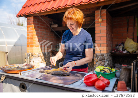 Woman seasoning fish and wrapping it in foil for outdoor grilling in her backyard. Homemade cooking, natural ingredients, and traditional barbecue preparation. 127678588