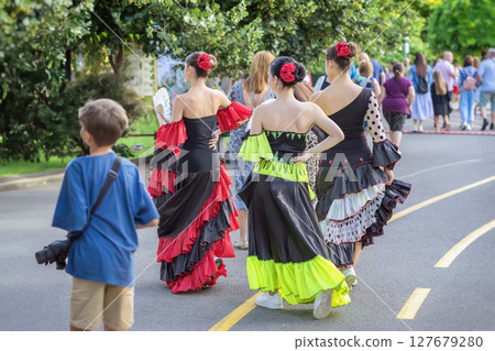 Folk ethnic flamenco dancers walking along street with people. Group of flamenco women dancers in bright dresses walking among crowd at cultural festival in park setting. Spanish Culture Festival 127679280