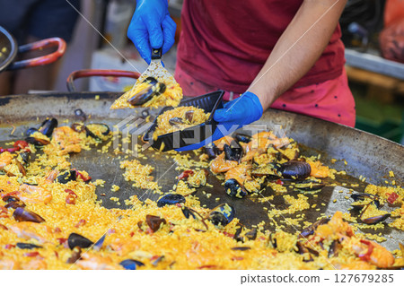 Chef in blue gloves serving seafood paella into plastic container from giant skillet during food event. Concept of paella, street food service. Spanish Culture and Food Festival 127679285