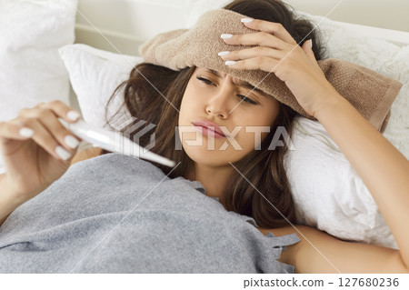 Sick young woman lying in bed under plaid, suffering from cold or flu and checking her temperature. Sick young woman lying in bed under plaid, suffering from cold or flu and checking her temperature. 127680236