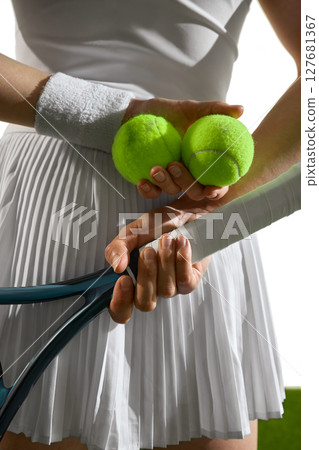 Female tennis player holding balls and racket with white sporty outfit and sunlight background 127681367