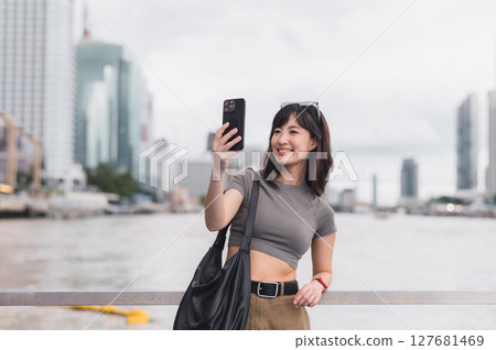 Solo Southeast Asian woman teenager blogger world travel. Local cultural tourism experience concept for explorer Bangkok Thailand. Express joy with smartphone, wait for friend boat Chao Phraya Solo Southeast Asian woman teenager blogger world travel. Local cultural tourism experience concept for explorer Bangkok Thailand. Express joy with smartphone, wait for friend boat Chao Phraya 127681469