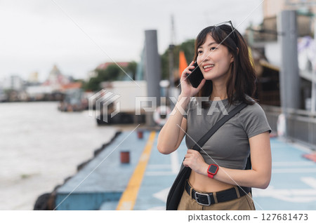 Happy solo Southeast Asian woman, teenager blogger, on world travel tourism experience in Thailand. She wait for boat on Chao Phraya River in Bangkok using smartphone to express joy to friend Happy solo Southeast Asian woman, teenager blogger, on world travel tourism experience in Thailand. She wait for boat on Chao Phraya River in Bangkok using smartphone to express joy to friend 127681473
