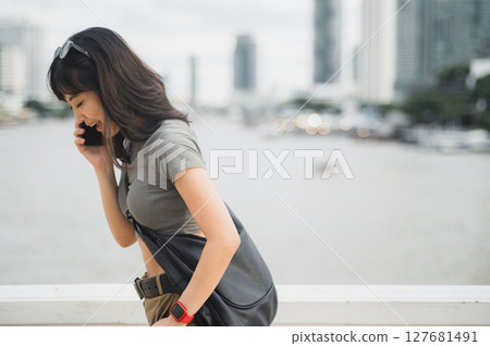 Solo southeast asian woman, teenager world travel explorer, on tourism trip in Asia. blogger has serious phone call with friend via smartphone by Chao Phraya River in Bangkok, Thailand 127681491