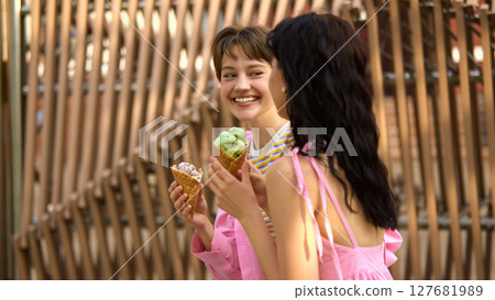 Two friends enjoying ice cream cones in outdoor urban setting with smiles and conversation Two friends enjoying ice cream cones in outdoor urban setting with smiles and conversation 127681989