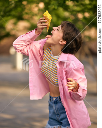 Melting moment. Young woman joyfully eating ice cream outdoors in pink shirt and denim shorts 127682031