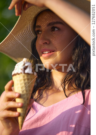 Styled and sweet. Woman enjoying ice cream cone under wide-brimmed hat on sunny day in outdoor setting 127682051