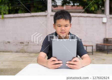 Thai teenage boy sitting at outdoor table using tablet wearing headphones 127682624
