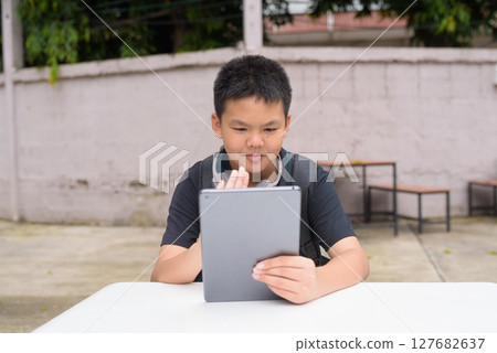 Thai teenage boy sitting at outdoor table using tablet wearing headphones 127682637