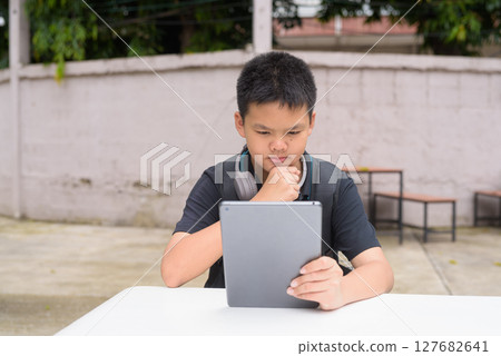 Thai teenage boy sitting at outdoor table using tablet wearing headphones Thai teenage boy sitting at outdoor table using tablet wearing headphones 127682641