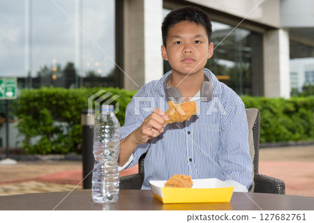 Thai boy eating fried chicken with headphones at outdoor table Thai boy eating fried chicken with headphones at outdoor table 127682761