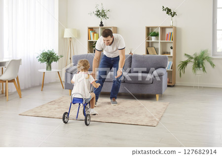 Cheerful father teaching his preschool daughter to ride bicycle, enjoying fun family time at home. 127682914