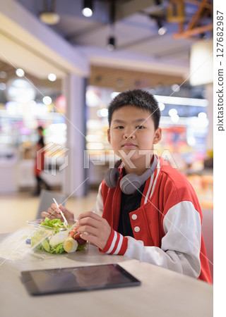 Teen Thai boy wearing headphones eating salad in cafeteria 127682987