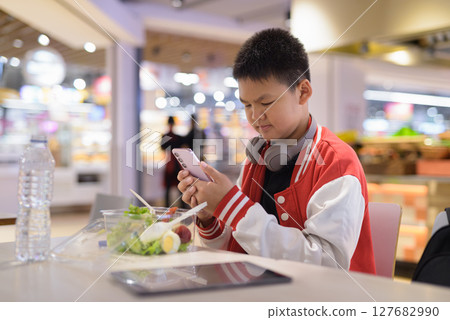 Teen Thai boy wearing headphones eating salad in cafeteria texting with phone 127682990