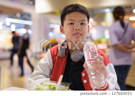 Thai teenage boy eating salad and drinking water indoors in cafe 127683033