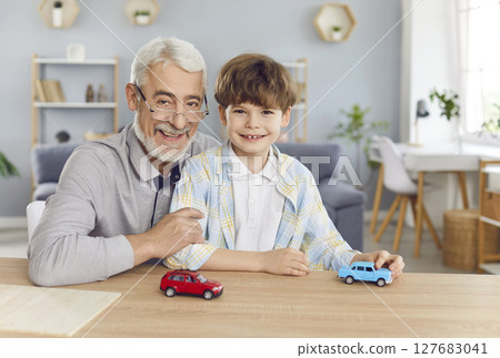 Grandfather with little cute boy grandson sitting at table at home and looking cheerful at camera. Grandfather with little cute boy grandson sitting at table at home and looking cheerful at camera. 127683041