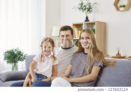 Cheerful family sitting on sofa with parents holding daughter on knees, making joyful home photo. Cheerful family sitting on sofa with parents holding daughter on knees, making joyful home photo. 127683124
