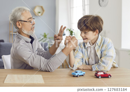 Grandfather arm wrestling with happy smiling boy grandson sitting at table at home together. Grandfather arm wrestling with happy smiling boy grandson sitting at table at home together. 127683184