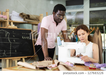Tailor observing seamstress assembling garment on sewing machine 127683226