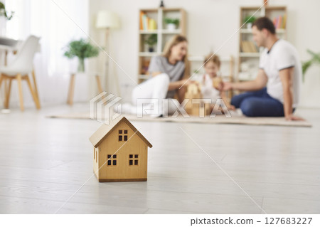 Small wooden toy house on floor at home, with happy parents and child playing in background 127683227