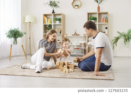 Joyful parents and their daughter playing with wooden blocks, building toy tower together at home. 127683235