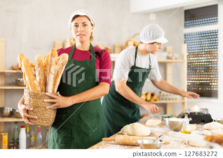 Portrait of positive female baker holding a basket of baguettes and fresh bread in hands 127683272