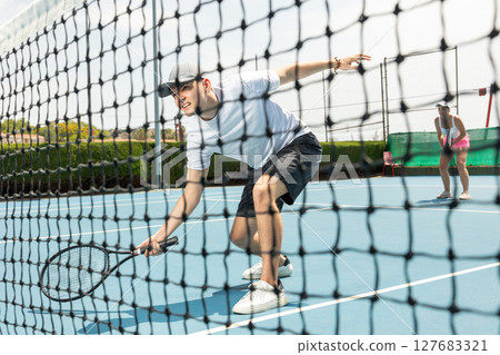 Portrait of man playing with male partner tennis doubles game on outdoors court. View through tennis net Portrait of man playing with male partner tennis doubles game on outdoors court. View through tennis net 127683321