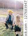 Little girl looks at her grandfather sitting on a stone on the lawn in a wreath of yellow leaves. Back view 127683947