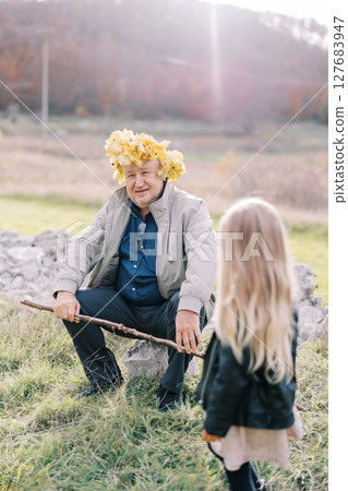 Little girl looks at her grandfather sitting on a stone on the lawn in a wreath of yellow leaves. Back view Little girl looks at her grandfather sitting on a stone on the lawn in a wreath of yellow leaves. Back view 127683947