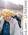 Little girl adjusts a wreath of yellow leaves on the head of a smiling grandfather sitting on the lawn 127683948