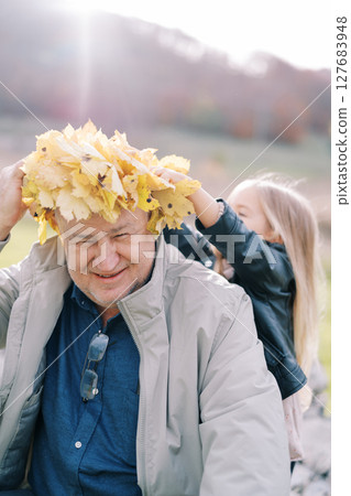 Little girl adjusts a wreath of yellow leaves on the head of a smiling grandfather sitting on the lawn Little girl adjusts a wreath of yellow leaves on the head of a smiling grandfather sitting on the lawn 127683948