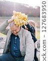 Little girl puts a wreath of autumn leaves on the head of her grandfather sitting on the lawn 127683950