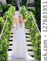 Bride stands on the steps of a hotel with an ivy-overgrown railing, touching her chin with her hands and looking down 127683951